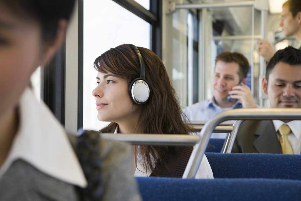 Commuters on a train with the focus on a dark-haired woman wearing headphones and looking out of the window. Using trains is a good way to create sustainable travel programs.