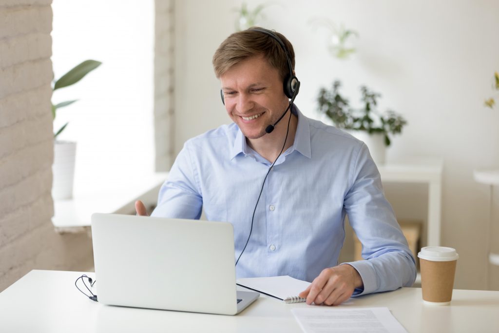 Smiling young businessman wearing headphones and looking at a computer screen with a takeaway coffee beside him on the desk planning sustainable travel programs